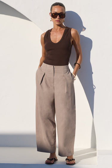 A model stands against a white architectural wall wearing a dark brown ribbed tank top, high-waisted wide-leg taupe trousers, and dark sunglasses.