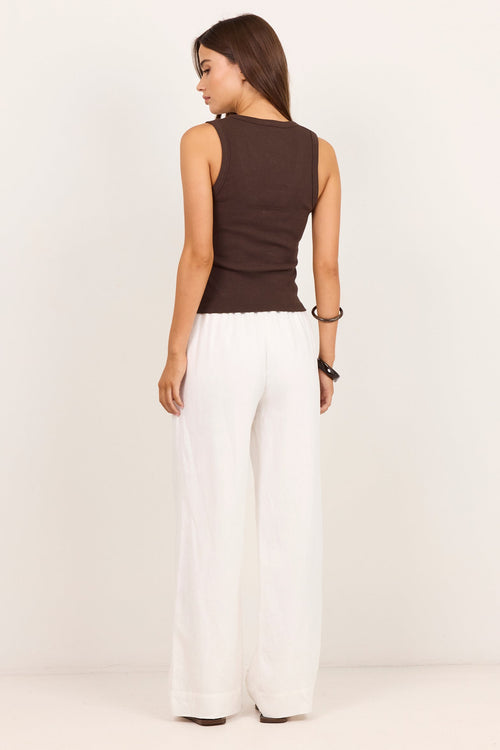 A woman is shown from the back wearing a sleeveless dark brown top and white wide-leg pants against a plain off-white studio background.