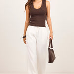 A woman modeling a dark brown ribbed tank top and white wide-leg drawstring pants, accessorized with sunglasses, a brown handbag, and black sandals, set against a plain off-white studio background.