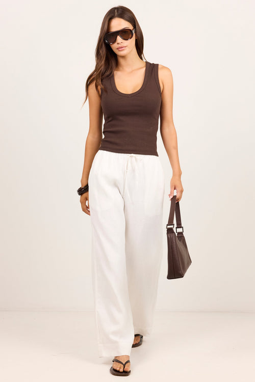 A woman modeling a dark brown ribbed tank top and white wide-leg drawstring pants, accessorized with sunglasses, a brown handbag, and black sandals, set against a plain off-white studio background.