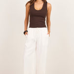 A woman modeling a dark brown ribbed tank top and white wide-leg linen pants against a clean, off-white studio background.