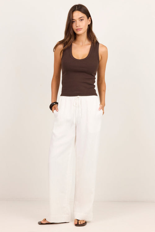 A woman modeling a dark brown ribbed tank top and white wide-leg linen pants against a clean, off-white studio background.