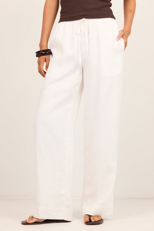 A model wearing white wide-leg drawstring pants, a dark brown ribbed tank top, and brown flat sandals, standing against a plain light beige studio background.