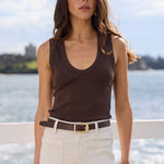 A woman stands outdoors by the water wearing a dark brown sleeveless scoop-neck tank top, cream-colored pants, and a dark belt with a gold buckle.