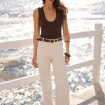 A woman stands on a sunny seaside walkway wearing a dark brown scoop-neck tank top, off-white wide-leg jeans with a thin black belt, and black sandals, with the ocean and white railing behind her.