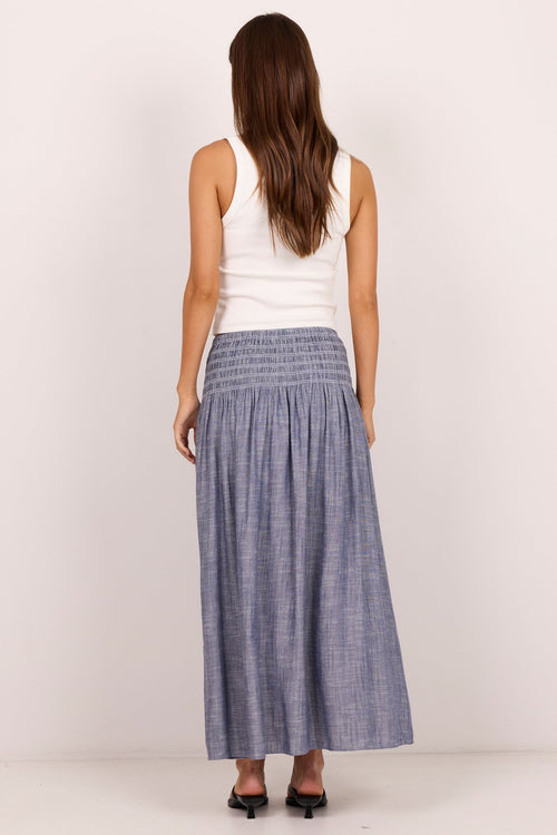 A woman viewed from behind wearing a sleeveless white top and a long, wide blue-gray pleated skirt, standing against a plain off-white studio background.