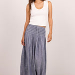 A woman modeling a pair of wide-leg, smocked blue-gray pants with a white ribbed tank top and black sandals, standing against a plain light gray studio background.