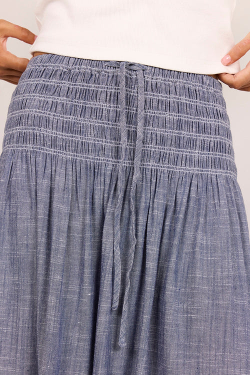 A close-up of a person wearing a smocked blue-gray maxi skirt with a drawstring waist, paired with a white top. The hands are holding the waistband to show its stretch.