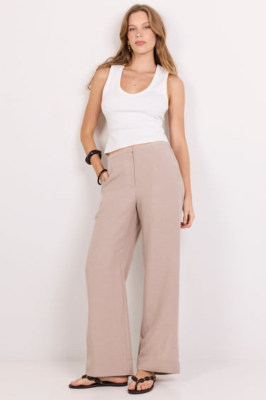 A woman modeling a pair of wide-leg taupe pants with a white tank top and black sandals, standing against a plain white studio background.