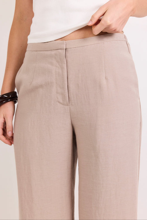 A close-up of a person wearing light beige wide-leg trousers and a white sleeveless top, with one hand lifting the hem of the top. The background is a clean, off-white studio setting.