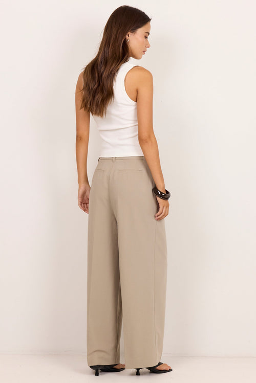 A woman stands in profile wearing wide-leg beige trousers, a white sleeveless top, and black sandals against a plain white studio background.