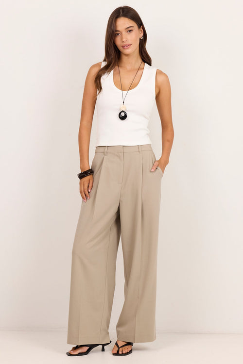 A woman modeling a pair of wide-leg beige trousers with a white sleeveless top, black sandals, and a long pendant necklace against a plain white studio background.