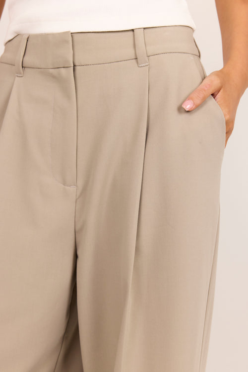 Close-up of a person wearing high-waisted, wide-leg trousers in a light taupe-beige color. The photo shows the waistband, front pleats, side pocket, and a hand with pink manicured nails resting in the pocket.