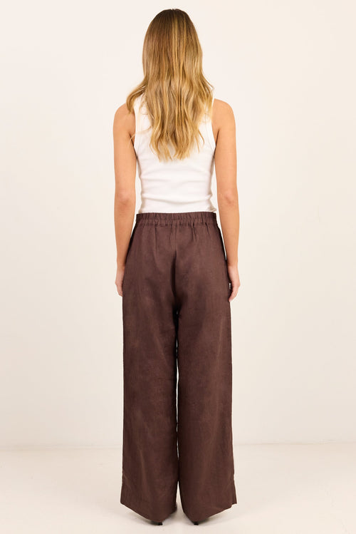 A woman viewed from the back wearing wide-leg brown pants and a white sleeveless top, standing against a plain off-white studio background.