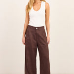 A woman modeling a pair of dark brown wide-leg trousers with a white sleeveless tank top, standing against a plain off-white studio background.