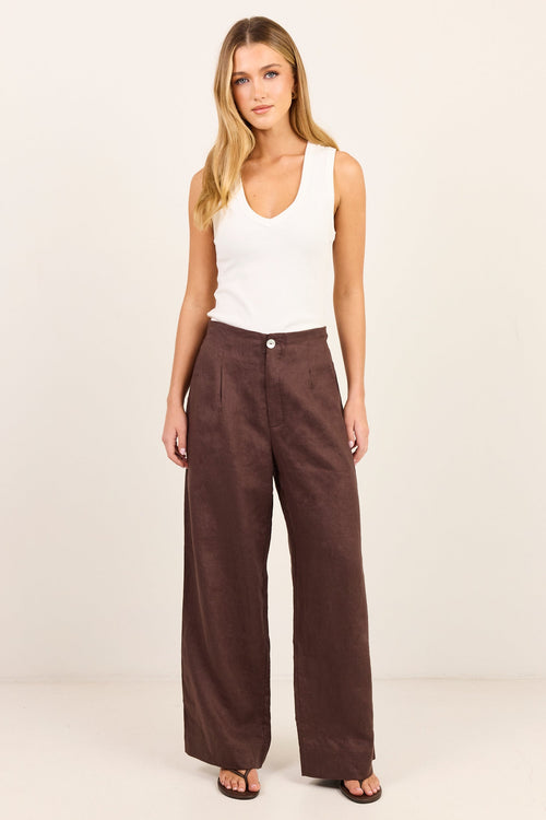 A woman modeling a pair of dark brown wide-leg trousers with a white sleeveless tank top, standing against a plain off-white studio background.
