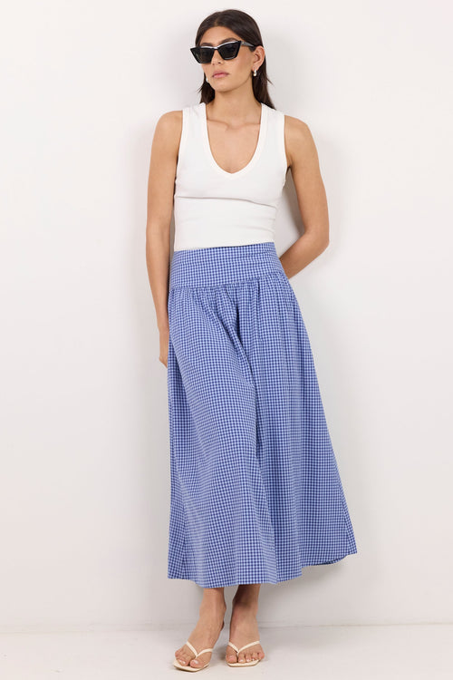 A woman modeling a white sleeveless top and a blue and white gingham pleated midi skirt, standing against a plain white studio wall.