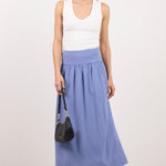 A model wearing a white sleeveless top and a blue and white gingham pleated maxi skirt, holding a small black handbag against a plain light gray studio background.