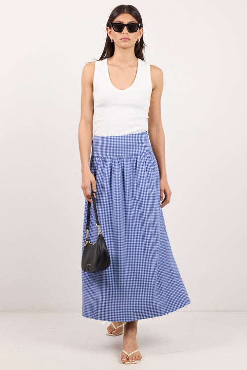 A model wearing a white sleeveless top and a blue and white gingham pleated maxi skirt, holding a small black handbag against a plain light gray studio background.