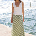 A woman stands on a concrete pier with the ocean in the background, wearing a white sleeveless top and a long green and white gingham maxi skirt with brown sandals.