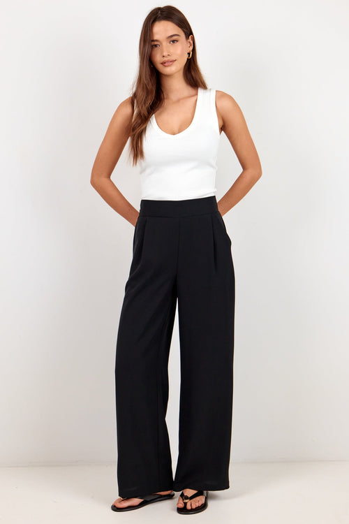 A woman modeling a white sleeveless tank top and black wide-leg trousers against a plain white studio background.