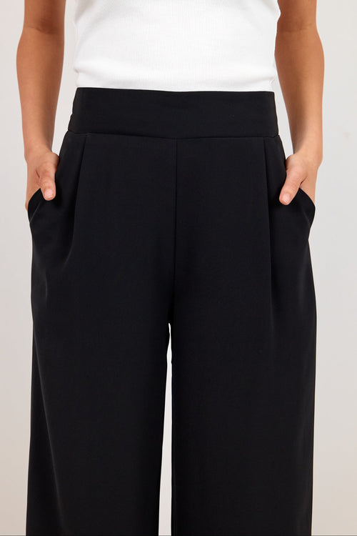 A model wearing black wide-leg trousers with side pockets and a white ribbed tank top, shown from the chest down against a light gray studio background.