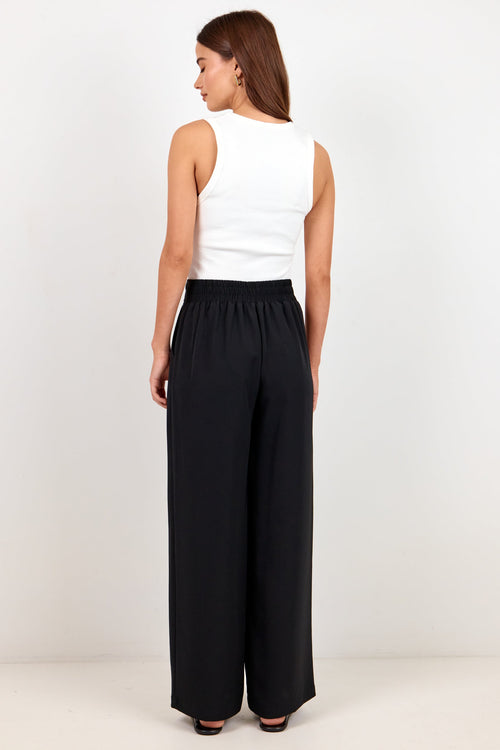 A woman shown from the back wearing a white sleeveless top and black wide-leg pants against a plain white studio background.
