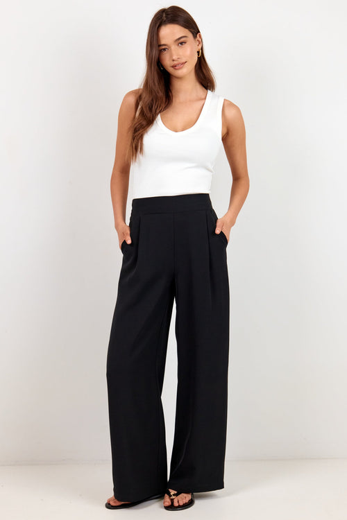 A woman modeling a white sleeveless tank top and black wide-leg trousers against a plain white studio background.