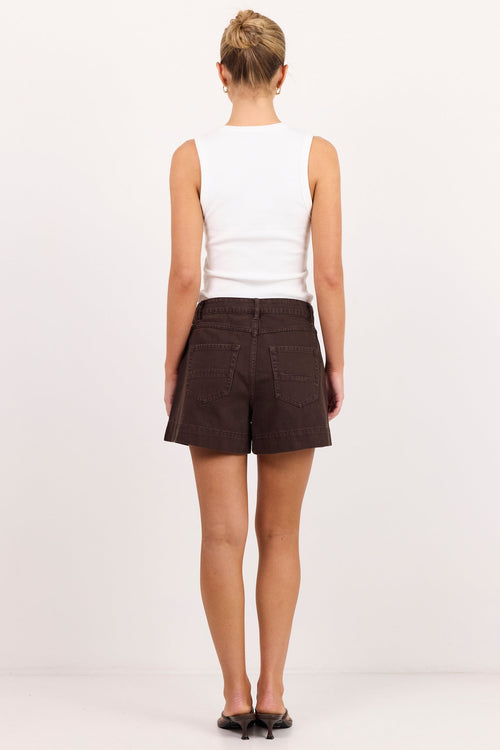 A woman viewed from behind wearing a white sleeveless top and dark brown shorts, standing against a plain white studio background.