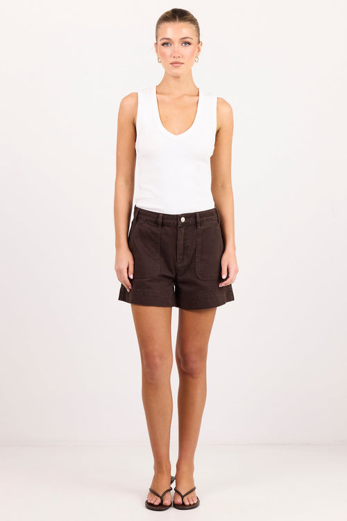 A woman stands against a plain white studio background wearing a white sleeveless tank top, dark brown shorts, and dark brown sandals.