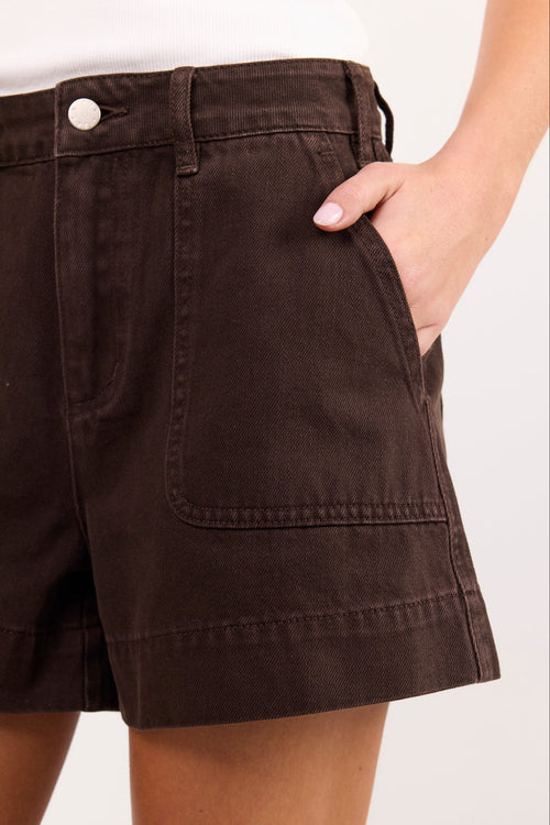 Close-up of a person wearing dark brown high-waisted shorts with front patch pockets and a white top, against a plain light background.