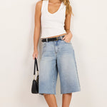 A woman modeling a pair of light-wash blue denim culotte jeans with a black belt, paired with a white tank top, black sunglasses, and black sandals, standing against a plain white studio background.