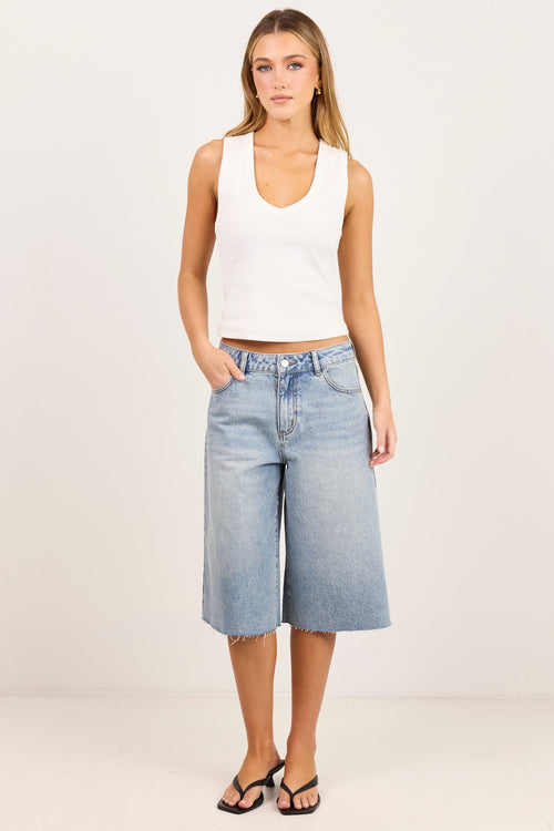 A woman modeling a white ribbed tank top and light-wash blue cropped denim shorts against a plain off-white studio background.
