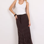 A woman modeling a long black maxi skirt with a white polka dot pattern, paired with a white sleeveless top and black strappy heels, against a plain white studio background.