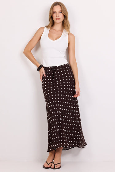 A woman modeling a long black maxi skirt with a white polka dot pattern, paired with a white sleeveless top and black strappy heels, against a plain white studio background.