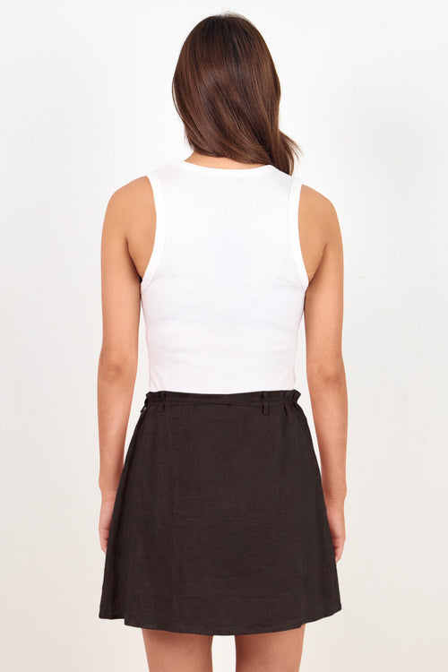 A woman shown from the back wearing a white sleeveless top and a dark brown flared skirt, standing against a plain off-white studio background.
