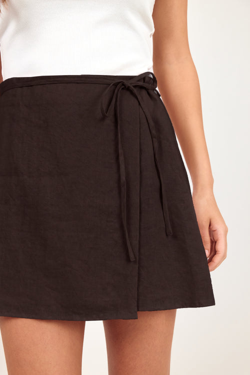 A close-up of a model wearing a dark brown wrap-style mini skirt with a tie waist, paired with a white sleeveless top, against a plain light background.
col_pattern
