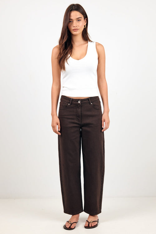 A woman modeling a pair of dark brown wide-leg jeans with a white tank top and black sandals, standing against a plain white studio background.