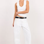 A model wearing a white sleeveless crop top, white wide-leg jeans, a black belt, and black sandals, standing against a plain white studio background.