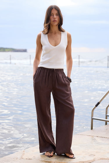 A woman stands on a stone pier by the water wearing a white sleeveless V-neck top and dark brown wide-leg pants, with a metal ladder visible to her right.