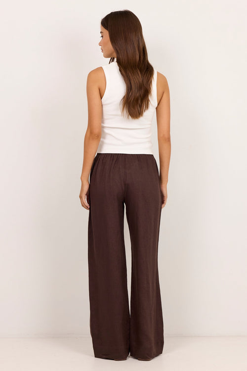 A woman is shown from the back wearing a white sleeveless top and dark brown wide-leg pants against a plain white studio background.