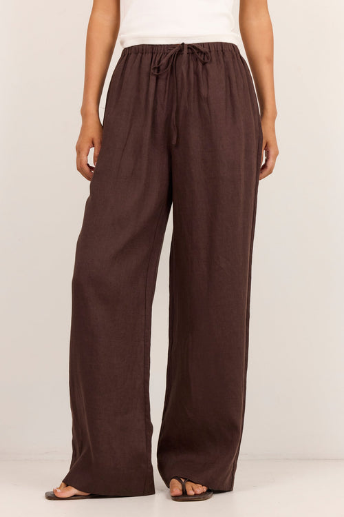 A model wearing a pair of dark brown wide-leg linen pants with a drawstring waist, paired with a white tank top and flat sandals, standing against a plain white studio background.