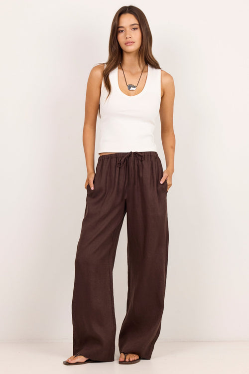 A woman modeling a pair of dark brown wide-leg drawstring pants with a white ribbed tank top, standing against a plain off-white studio background.