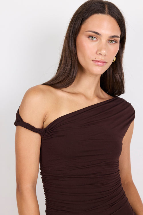 A woman wearing a dark brown off-the-shoulder ruched top, shown from the chest up against a light gray studio background.