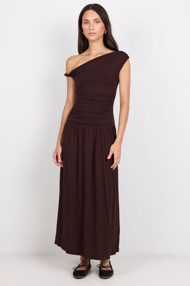 A woman modeling a dark brown one-shoulder midi dress with a gathered bodice and flowing skirt, standing against a plain white studio background.