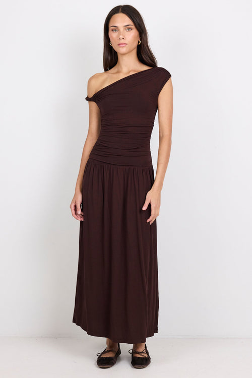A woman modeling a dark brown one-shoulder midi dress with a gathered bodice and flowing skirt, standing against a plain white studio background.