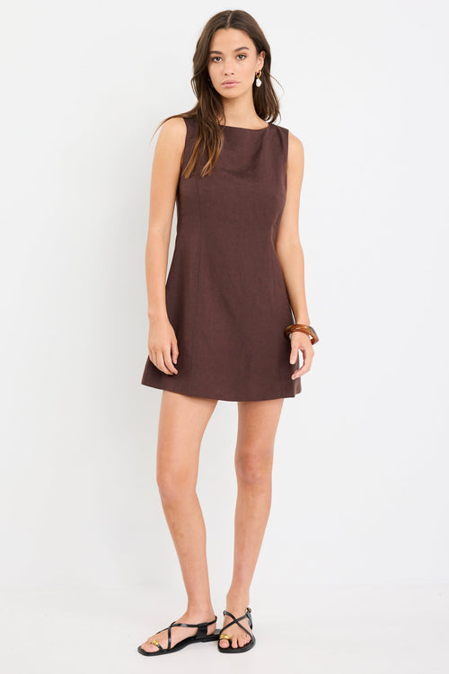 Woman wearing a sleeveless chocolate brown mini dress with a boat neckline and fitted silhouette, styled with black sandals.