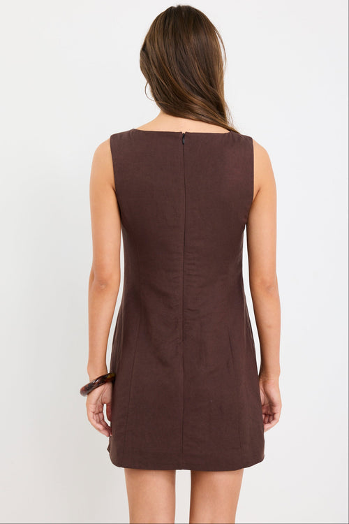 Woman wearing a sleeveless chocolate brown mini dress with a boat neckline and fitted silhouette, styled with black sandals.