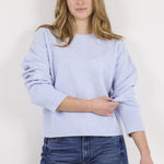 A woman modeling a light periwinkle blue knit sweater with wide three-quarter sleeves and a ribbed hem, paired with dark blue wide-leg jeans, against a plain off-white background.