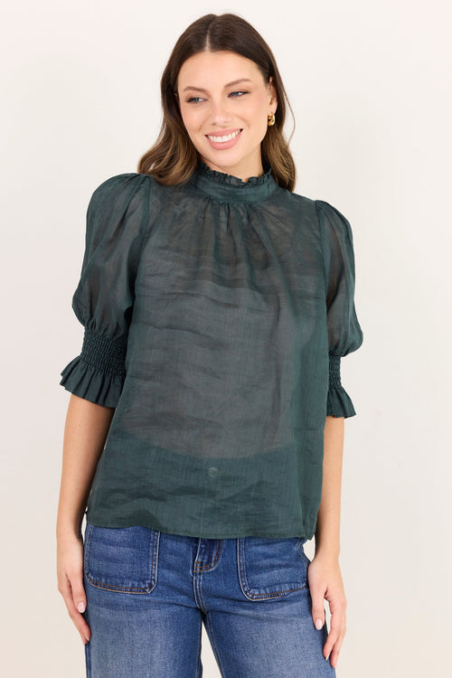 A woman wearing a dark green sheer puff-sleeve blouse with a ruffled high neckline, paired with blue jeans, against a plain off-white background.